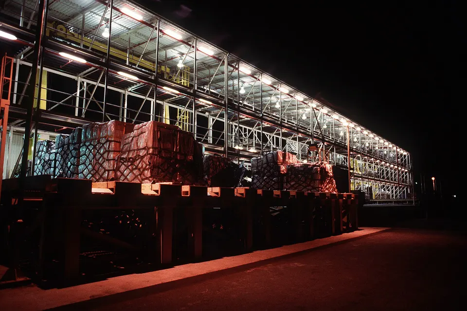 Pallets wrapped in red plastic sit on a raised metal rack beside a well-lit, multi-level steel structure at night.