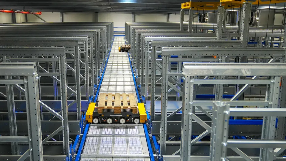 Long aisles of galvanized steel pallet racking in a warehouse, with a yellow automated vehicle carrying a wooden pallet on a central track.