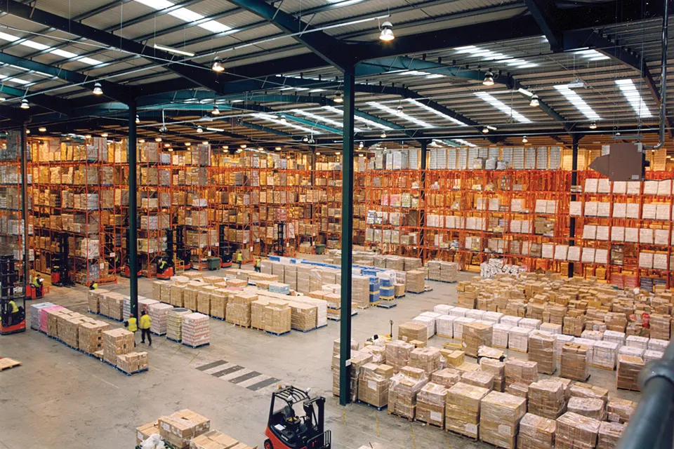 Large warehouse interior with tall orange pallet racking filled with boxes, stacked pallets on the floor, and a red forklift in the foreground.