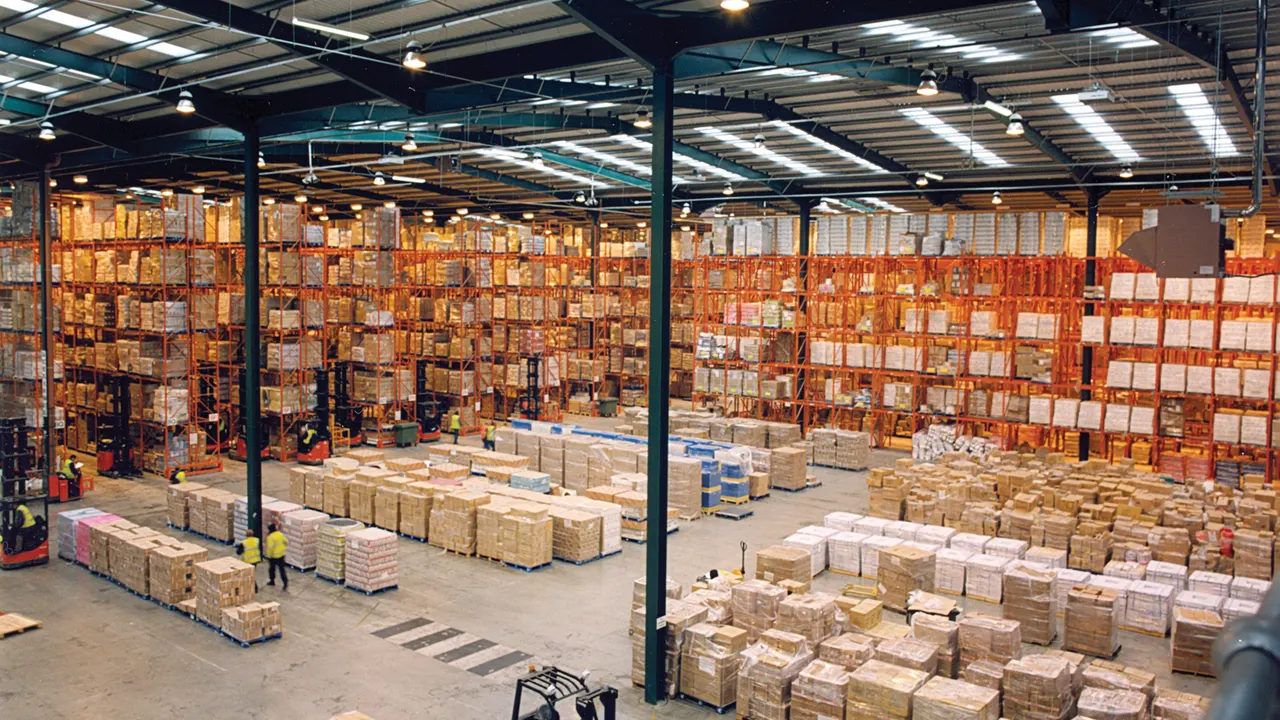 Large warehouse interior with tall orange pallet racking filled with boxes, stacked pallets on the floor, and a red forklift in the foreground.