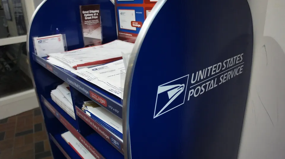 A blue USPS shipping supplies display holds envelopes and mailers, with a sign reading 'Great Shipping Options at a Great Price' and the United States Postal Service logo.